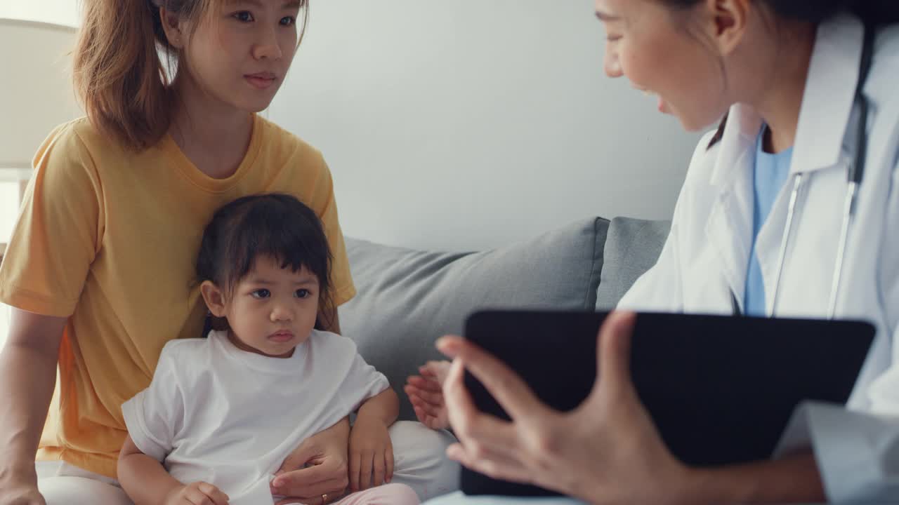 年轻的亚洲女儿科医生和小女孩病人使用数字平板电脑分享良好的健康测试消息与快乐的妈妈坐在沙发在家里。