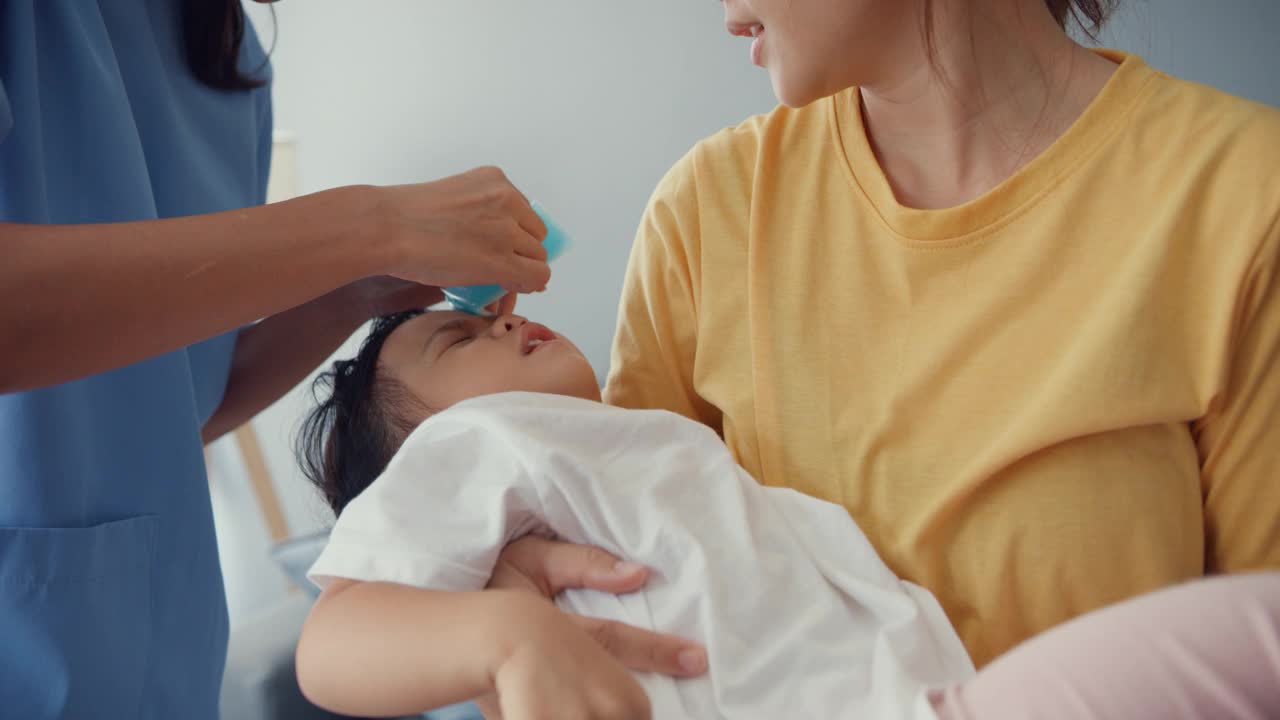 亚细亚女儿科医生将退热凝胶贴在婴儿额头上，患者在家中客厅与母亲就诊。