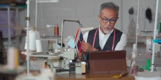 亚洲华人高级男子在他的工作室展示变化纺织品织物样本目录视频电话客户与数字平板电脑