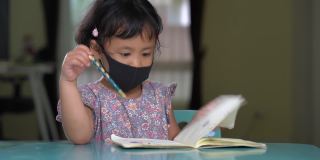 亚洲小女孩戴着防护口罩，在餐桌上专心读书。
