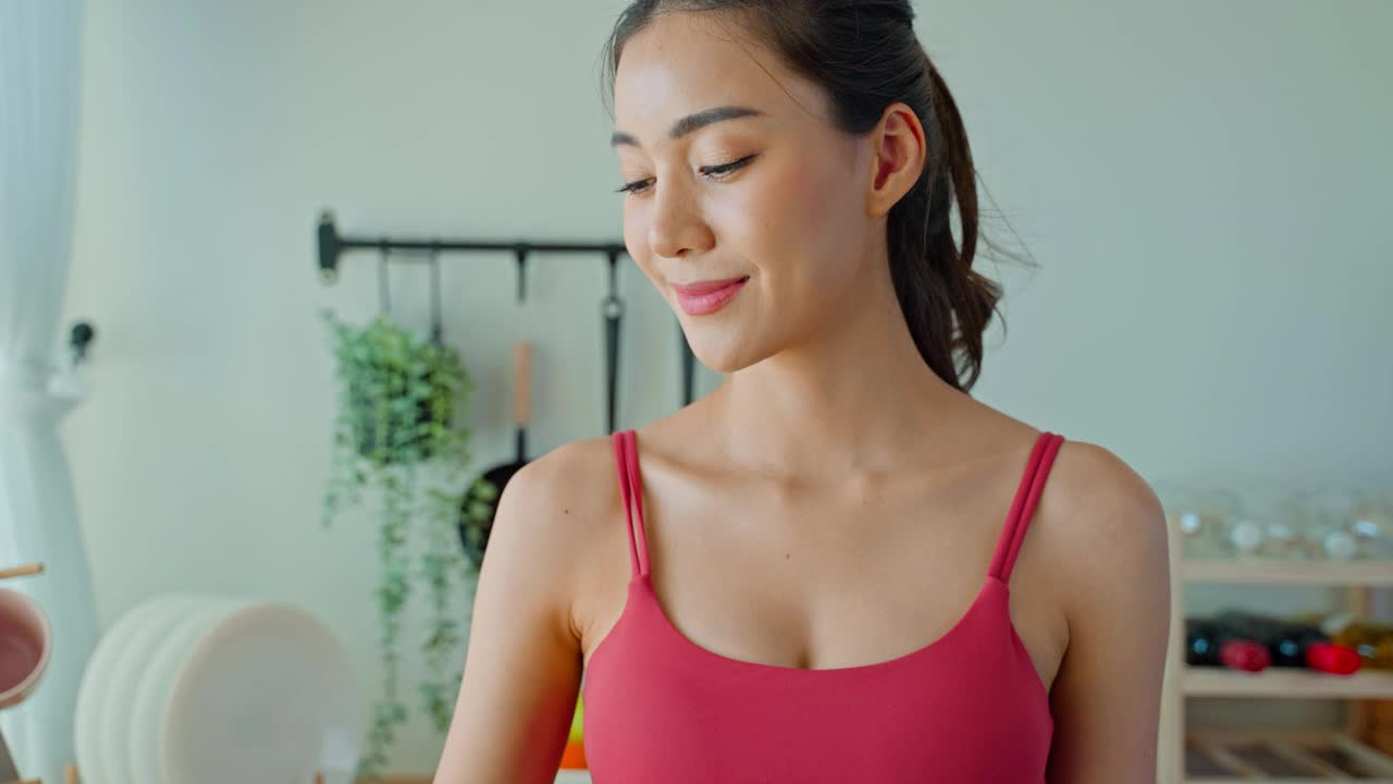 亚洲美女肖像在家里锻炼后喝水。年轻美丽口渴的运动女孩在家里的厨房为保健锻炼后喝一杯干净的天然矿物质
