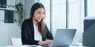 亚洲商务女性呼叫中心正装工作在工作场所。漂亮漂亮的职业女性电话销售人员坐在桌子上，戴着耳机和客户在办公室交谈