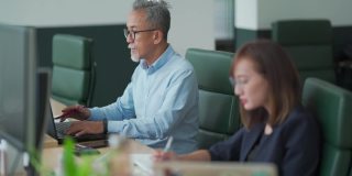 亚洲华人工作高级男子系使用笔记本电脑与他的同事在开放式办公室工作