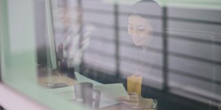 透过窗户，一位亚洲华人女性专注于阅读文献研究，与同事并肩工作在工作站