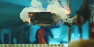 专业厨师烹饪火焰风格。用明火在平底锅中准备菜肉。厨师在商业厨房的煤气灶上煎拌食物。餐馆里，一名男子正在用火红的平底锅煎牛排。