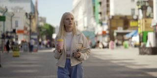 一位有创意的时尚女性，一头浅卷发，手里拿着书和平板电脑，充满热情地走在街上。她用一个漂亮的粉红色杯子喝咖啡，环顾四周，欣赏这座城市。