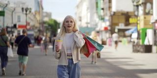 在大城市的精品店拿着包的时髦女人。她背着一堆彩色的袋子走在步行街上。她手里拿着一个原装的杯子，里面装着美味的咖啡。时装店和服装店。