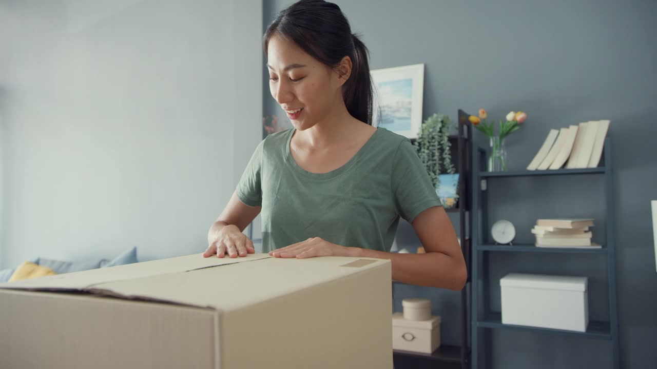 快乐美丽的亚洲女士打开纸箱从网上市场快递包裹在家里的客厅。
