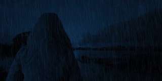 一个戴着兜帽的人站在雨与闪电中，在雷雨中拍摄了一个湖。