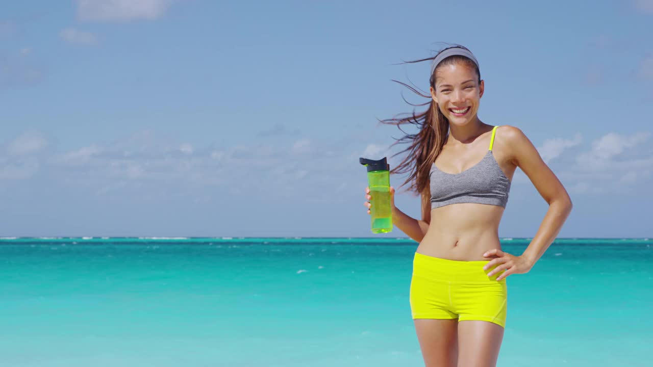 运动员在户外沙滩上进行健身跑步后饮用瓶装水。口渴的女人喝着冷饮，在户外剧烈运动后满头大汗竖起大拇指。缓慢的运动。