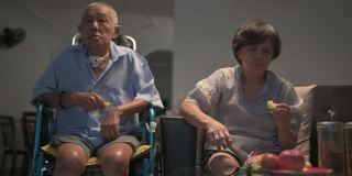 亚洲华人老夫妇坐在轮椅上享受电影之夜，坐在客厅的沙发上看电视