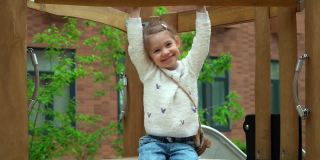 小女孩的肖像坐在操场上的滑梯上，看着相机，在绿色的春天公园里笑着。快乐的孩子在户外玩耍。