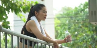 美丽的亚洲健身女人穿着运动服和毛巾在跑步后喝水休息。疲惫的女性在户外公园训练后休息