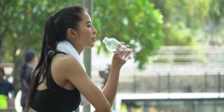 美丽的亚洲健身女人穿着运动服和毛巾在跑步后喝水休息。疲惫的女性在户外公园训练后休息
