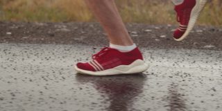 男子铁人三项运动员的腿穿着红色运动鞋在下雨时在沥青上奔跑，侧视图，特写。运动员在雷雨中沿路奔跑