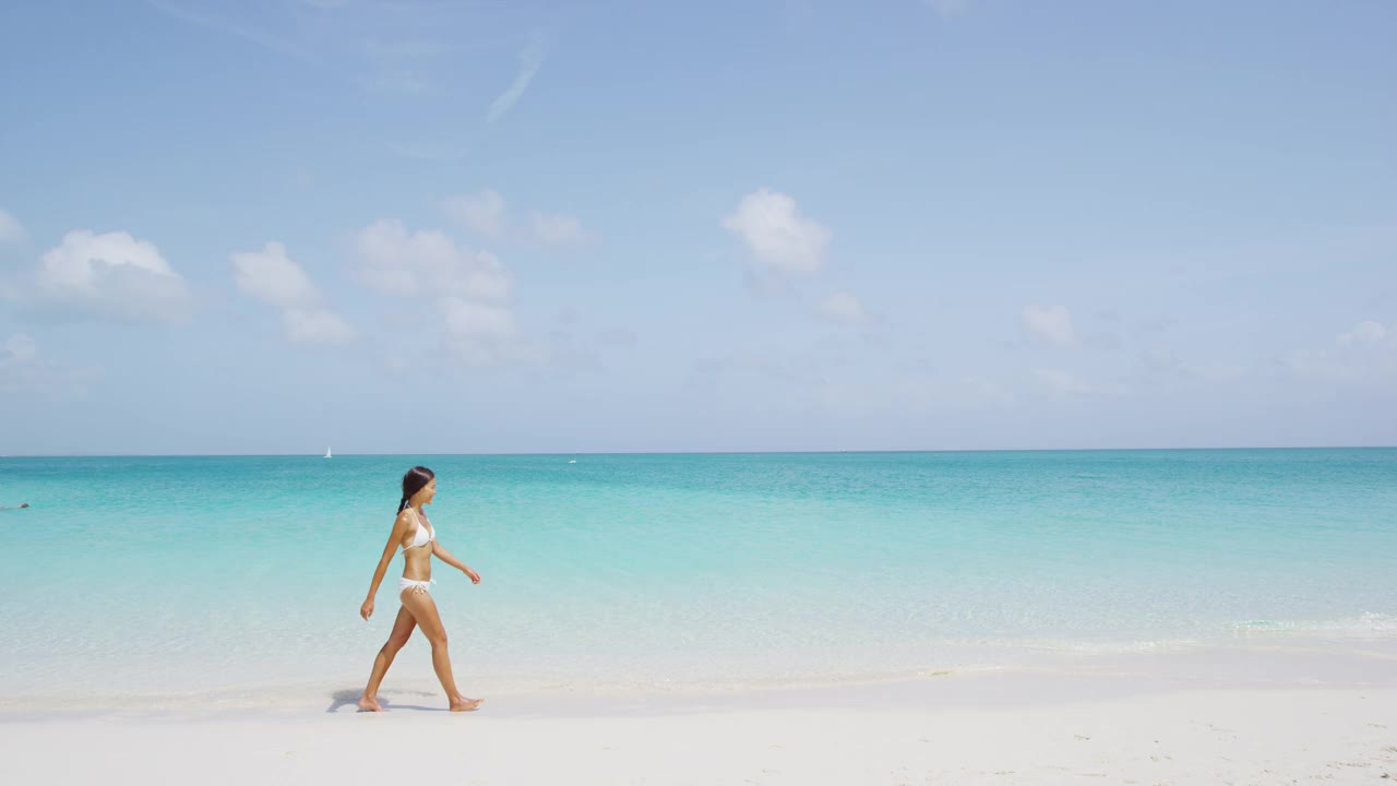 穿着白色比基尼的美女在海滩上散步。年轻的女性在暑假期间享受大自然。湛蓝天空下的海景。