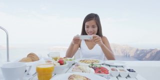 女人用手机app拍早餐照片。女孩在奢华旅行度假时拍美食照片，用于社交媒体。欧洲希腊圣托里尼度假胜地的美女。