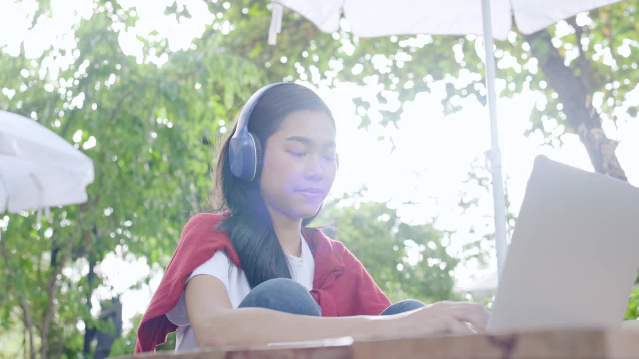 亚洲美丽的少女戴着耳机使用笔记本电脑坐在花园里学习在线。视频网络研讨会。在家远程视频会议。业务网上购物。在家里工作。网上注册