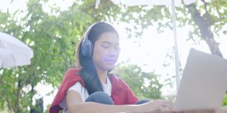 亚洲美丽的少女戴着耳机使用笔记本电脑坐在花园里学习在线。视频网络研讨会。在家远程视频会议。业务网上购物。在家里工作。网上注册