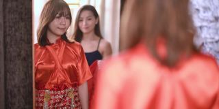 在婚纱店，亚洲的中国新娘在姐姐的帮助下，一边看着镜子一边试穿中国传统的婚纱
