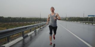 亚洲体育三项全能运动员在下雨时在高速公路上跑马拉松，正面视图。体格健壮的男人沿着公路跑步做有氧运动