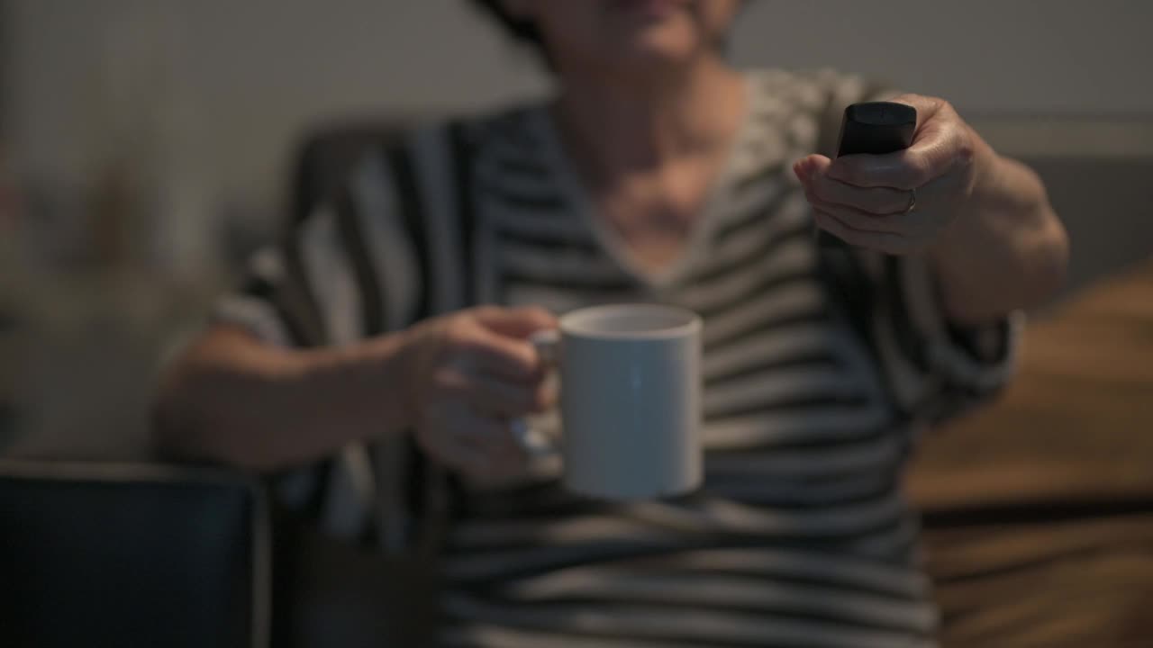 亚洲华人老年妇女手握电视遥控器，选择频道，在周末的闲暇时间独自一人在家看电视