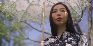 自信的年轻亚洲女性肖像，站在夏日公园，撑着太阳伞，环顾四周，享受大自然的美丽。美丽苗条的千禧一代在阳光明媚的春天花园里休息。