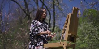 美丽苗条的亚洲女人在和服喝茶站在画架在阳光明媚的春天公园户外。这是一位才华横溢的女画家的肖像。美丽的概念。