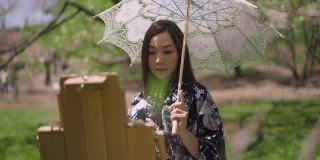 迷人的微笑自信的亚洲画家画在春天阳光明媚的公园微笑。美丽的日本年轻女子的肖像，穿着和服，手持白色太阳伞，享受户外慢动作。