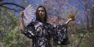 年轻苗条的日本女子手拿扇子慢动作跳舞。美丽的亚洲舞者穿着和服在公园里享受阳光明媚的春天。