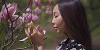 在阳光明媚的春天公园里，快乐美丽的亚洲女人在树上享受粉红色樱花的香味。美丽的日本千禧一代穿着和服在阳光下闻花瓣的慢动作。