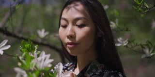 自信美丽的亚洲年轻女子在阳光下盛开的白花树前摆姿势的头像。轻松美丽快乐的女士站在春天公园的户外看着相机。