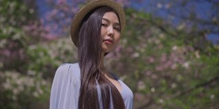一个自信的亚洲女子，穿着蓝色上衣，手拿草帽，站在盛开的樱花公园，在阳光明媚的春日里看着镜头。优雅美丽的日本女士在花园里摆姿势的肖像。