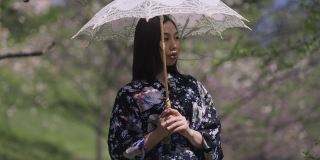 迷人的亚洲美女肖像，手持白色太阳伞，站在春天公园转向相机微笑。自信迷人的日本女士穿着和服在户外摆姿势。