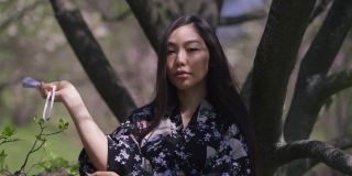 自信的年轻日本女子的肖像，穿着和服，打开风扇看着相机站在阳光下的树。美丽苗条迷人的女士在春天夏天公园摆姿势的正面视图。