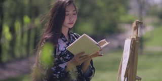 微笑美丽的亚洲女人在阳光下看书与模糊的画架在背景。照片中，日本千禧一代穿着和服，在阳光明媚的春天公园里享受着乐趣。