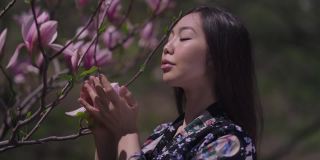 年轻美丽的亚洲女子欣赏着树枝上粉红色的樱花，看着镜头抚摸着脸。自信迷人的美女肖像穿着和服在阳光下摆姿势户外。