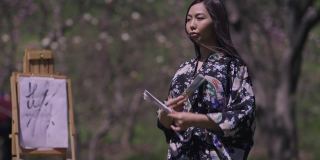 美丽的亚洲年轻女子穿着和服在春天的樱花花园与扇子跳舞微笑与画布画在背景模糊的画架。在阳光明媚的日子里，自信的日本女士的肖像。