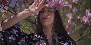 在春天公园里，一位自信的亚洲年轻女子身穿和服，在盛开着粉红色花朵的樱花前摆姿势。自信美丽的日本女士在户外休息，看着相机。
