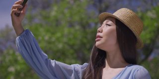 快乐的亚洲女人在春天夏天公园的阳光下，站在智能手机聊天。这是一位戴着草帽、欢快、轻松、优雅的千禧一代在使用手机视频聊天应用。旅游和生活方式。