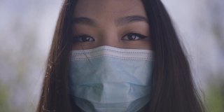 一名戴着新型冠状病毒口罩的亚洲女性站在公园室外看着摄像机的照片。这是一位棕色眼睛、黑头发的美丽女士在冠状病毒大流行期间的特写照片。
