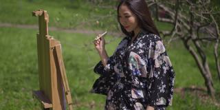 旁边的亚洲女人穿着和服，用毛笔在画布上微笑。这是一幅灵感迸发、技艺精湛的日本画家在户外享受阳光明媚的春日的肖像。缓慢的运动。