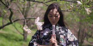 年轻自信的亚洲美女正面肖像，手握扇子看着相机。美丽的日本女士在春天的公园里与盛开的树木合影。缓慢的运动。