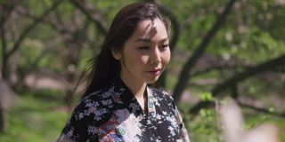 快乐自信的亚洲年轻女子穿着和服站在春天公园的阳光下微笑着看着相机。愉快放松的日本女士肖像在阳光下户外慢动作。