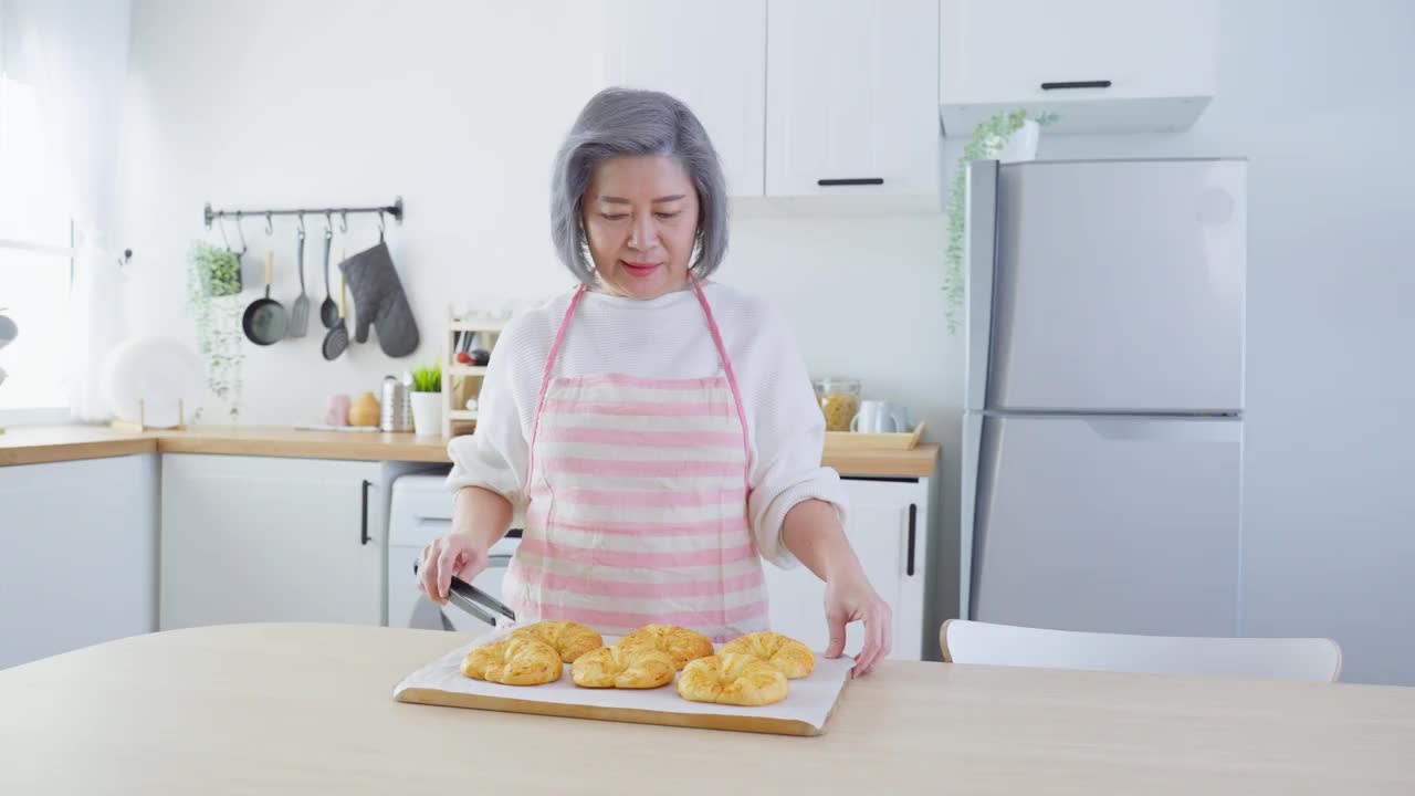 亚洲可爱的家庭，年轻的女儿看着老妈妈在厨房做饭。美丽的女性享受打发闲暇时光，抱着年迈的妈妈在家里的餐桌上烤牛角面包。活动的关系