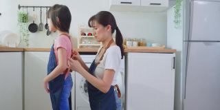 亚洲慈爱的妈妈在打扫房子前为年轻的女孩孩子系上围裙。爱美丽的父母妈妈帮助和支持可爱的小女儿在家里的厨房穿围裙。活动关系的概念。