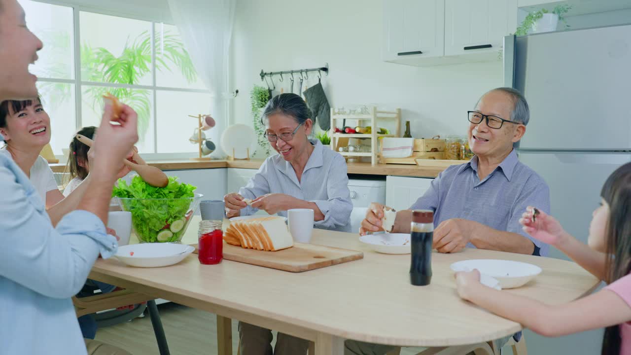 亚洲大家庭在家里的餐桌上一起吃午饭。年长的爷爷奶奶、年轻的夫妇和年幼的女儿感到高兴，在家里享受吃面包。活动关系的概念