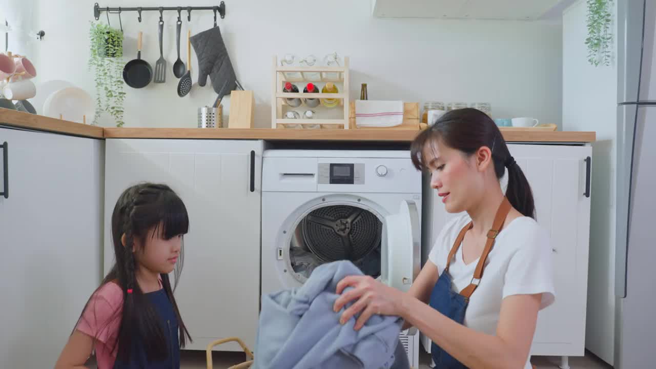 亚喜一家人待在家里，妈妈教女儿洗脏衣服。年幼可爱的小女孩帮助父母妈妈把衣服放在家里的洗衣机里。Domestic-Housekeeping。