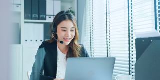 亚洲商务女性呼叫中心正装工作在工作场所。漂亮漂亮的职业女性电话销售人员坐在桌子上，戴着耳机和客户在办公室交谈