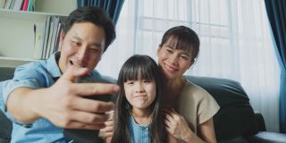 亚洲父母带着小女儿与家人在线视频通话。慈爱的爸爸、妈妈和小女孩坐在客厅的沙发上，周末在家享受用手机和亲戚聊天的乐趣。